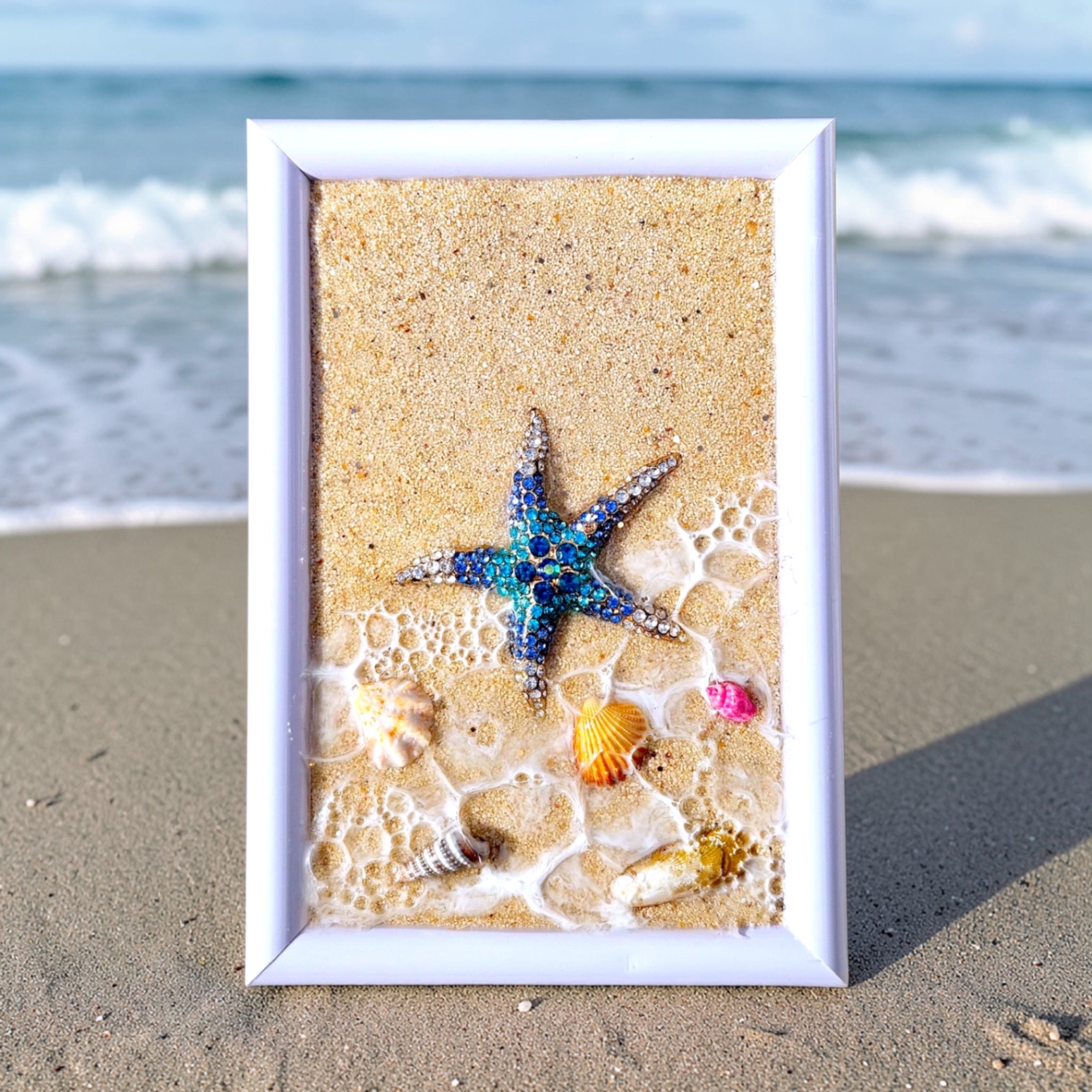 White frame featuring a blue jeweled starfish on sand with resin surf and shells.